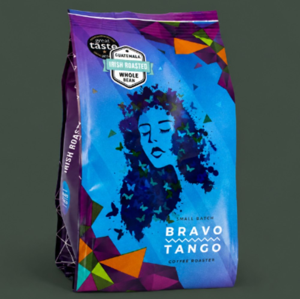 A bag of Bravo Tango Coffee Roast whole bean coffee, featuring a vibrant blue and purple design with a stylized graphic of a woman's face made of dark blue leaves, set against a dark gray background.