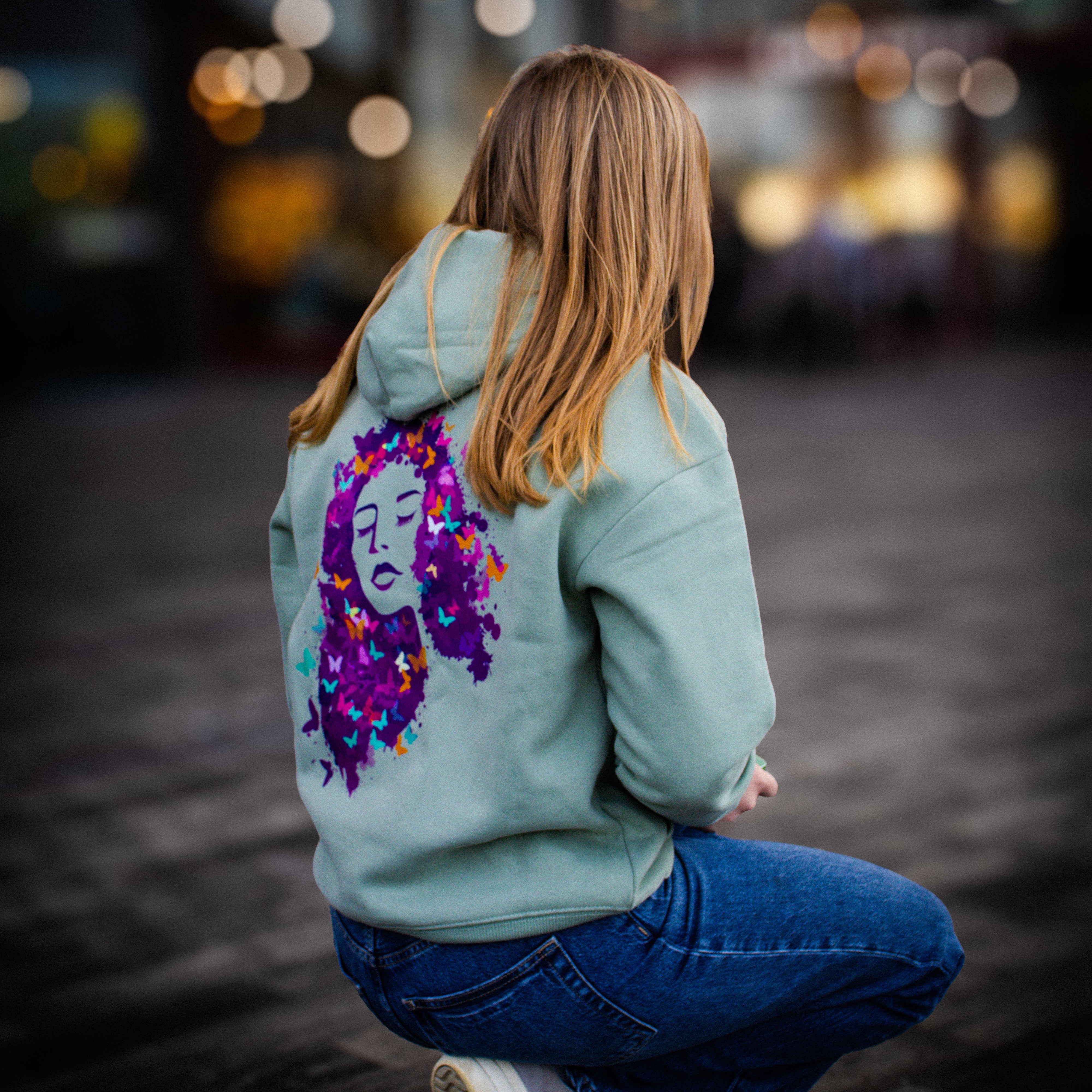 Person wearing a light blue hoodie with a colorful design on the back, sitting on a dark surface with blurred lights in the background.